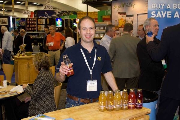 A smiling man with an exhibitor badge holds a Honeydrop bottle beside a row of Honeydrop drinks on a wooden counter. Attendees mill about on the show floor behind him, near a Sage Valley sign.