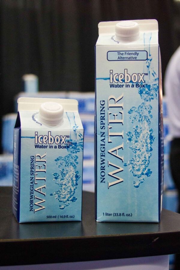 Two Icebox Norwegian Spring Water cartons on a dark table: a 500 ml Icebox Jr. carton and a taller 1 liter carton, with more blue cartons blurred behind them.