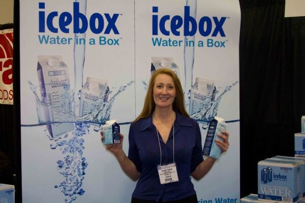 A smiling woman with long red-blond hair and an exhibitor badge holds up two small Icebox water cartons. Icebox 'Water in a Box' banners are behind her and boxes of water are at right.