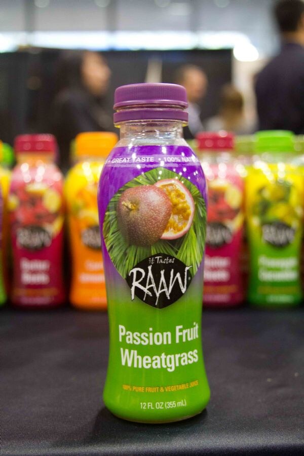 A 12 oz bottle of It Tastes Raaw Passion Fruit Wheatgrass juice with a purple cap stands on a black table in front of other blurred Raaw juice bottles.