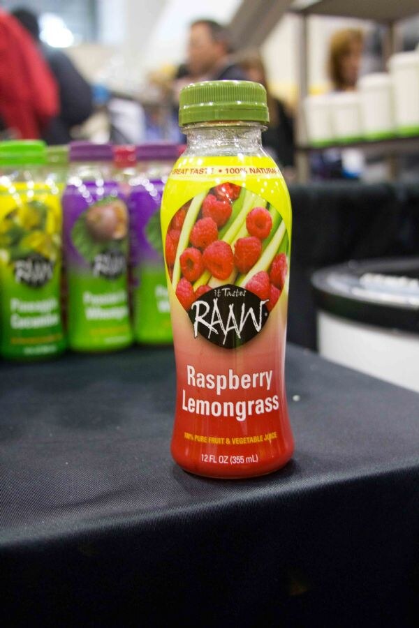 A 12 oz bottle of It Tastes Raaw Raspberry Lemongrass juice with a green cap sits on a black-draped table, with other Raaw bottles blurred behind it.