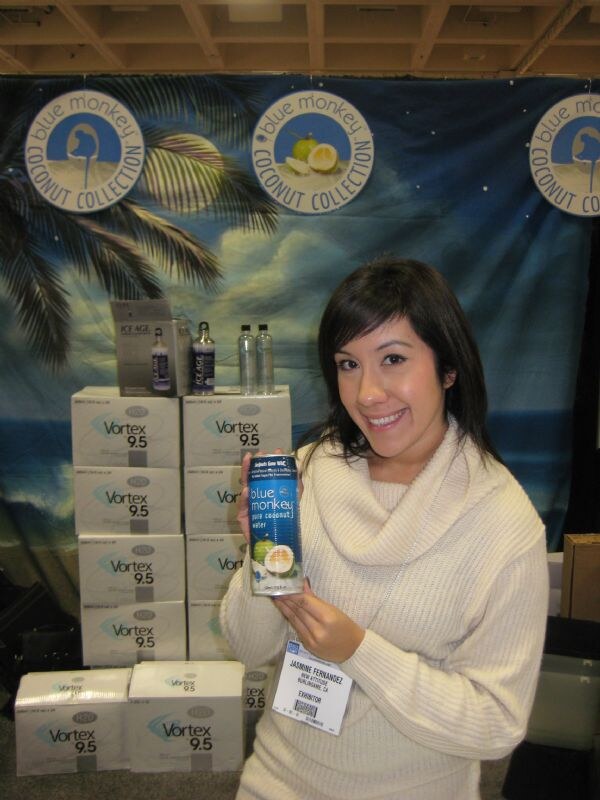 Jasmine Fernandez smiles and holds a can of Blue Monkey pure coconut water. Stacked Vortex 9.5 cases and a Blue Monkey Coconut Collection backdrop are behind her.