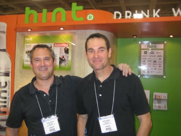 Two men in black polo shirts with exhibitor badges stand side by side in front of the green and orange Hint booth. Keith Busby of Hint, Inc. is on the right; a Hint bottle is at left.