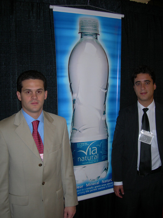 Two men in suits stand on either side of a tall Via Natural banner showing a water bottle, in front of a dark curtain.