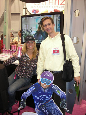 A woman in a cap holding a pink can and a man in a pale shirt with a shoulder bag stand behind a cardboard cutout of a skier in a Chevy Truck and Visa suit, near a screen.