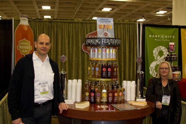 A man and a woman stand at a round display of Kombucha Wonder Drink bottles and cups; a Gary & Kit's Napa Valley banner is at right and a large bottle graphic at left.