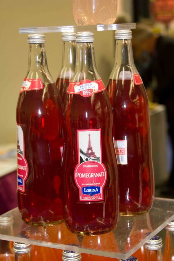 Several glass bottles of Lorina Sparkling Pomegranate premium French soda with red liquid and Eiffel Tower labels sit on a clear acrylic display stand.