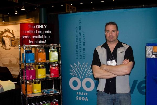 A man with arms crossed stands beside a wire rack of Oogave agave soda bottles in front of a teal Oogave Agave Soda banner.