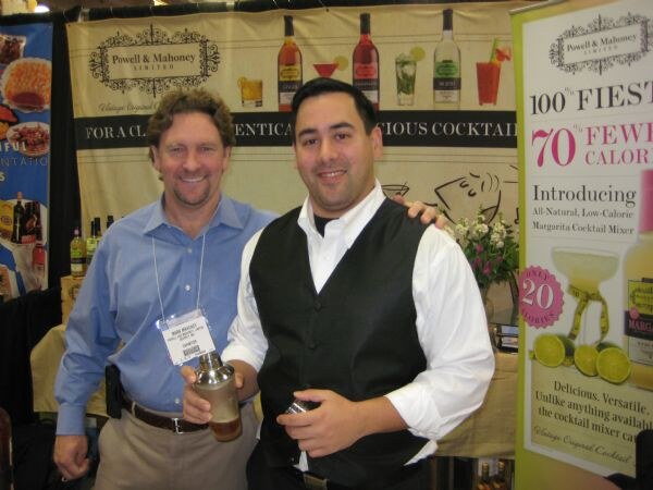 A man in a blue shirt and a man in a black vest holding a cocktail shaker smile in front of Powell & Mahoney banners advertising cocktail mixers.