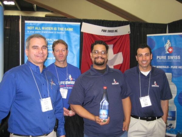 Four men in blue Pure Swiss shirts and badges stand in a booth in front of a Swiss flag and Pure Swiss banners. The man second from right holds a bottle of Pure Swiss water.