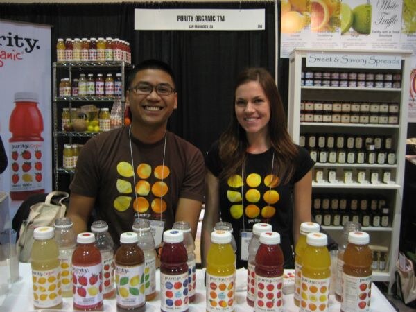A smiling man and woman in matching brown and black shirts with orange slice graphics stand behind a table of Purity Organic juice bottles, with a Purity Organic sign and shelves behind them.