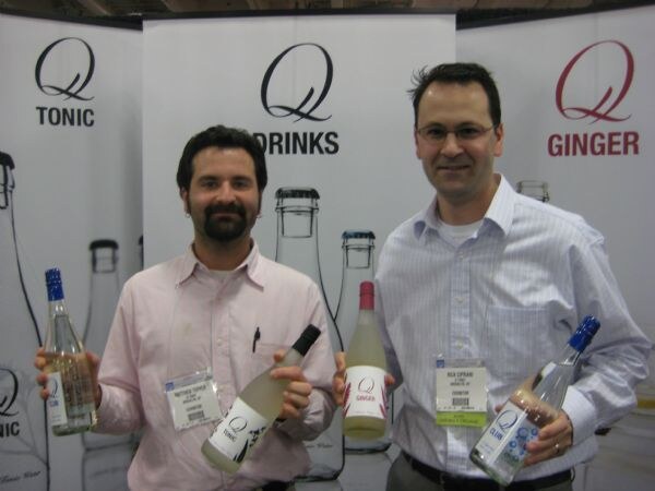 Two men with name badges hold bottles of Q Tonic and Q Ginger in front of banners reading Q Tonic, Q Drinks and Q Ginger. The man on the left has a beard; the man on the right wears glasses.