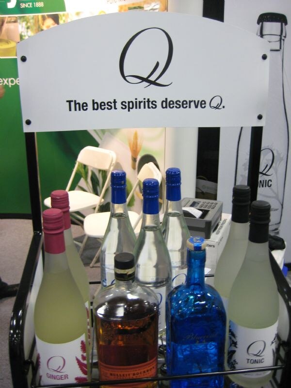 A black display rack under a sign reading "The best spirits deserve Q." holds bottles of Q Ginger and Q Tonic, a bottle of Bulleit Bourbon and a blue glass bottle.