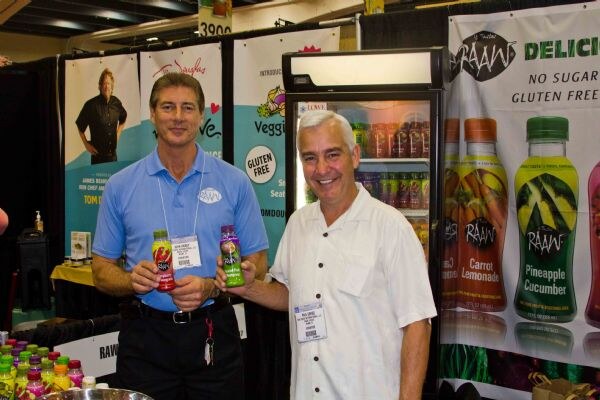 Two men with name badges stand in a RAAW booth; the man on the left holds two small bottles. Behind them are a refrigerated case and banners for RAAW Carrot Lemonade and Pineapple Cucumber.