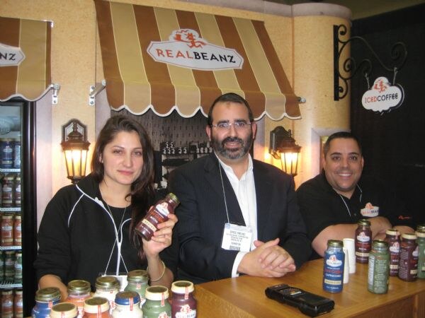 A woman holding a bottle of iced coffee, a bearded man in a suit and a smiling man in a black shirt stand behind a counter under a Real Beanz awning, with bottled drinks on the counter.