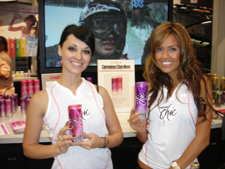 Two smiling women in white Chic tank tops each hold a pink or purple can of Chic, with more cans on a counter and a screen behind them.