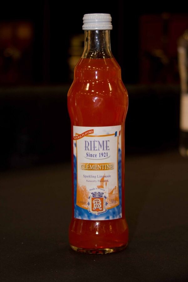 A bottle of Rième Clémentine sparkling limonade with an orange liquid and a white cap, labeled Since 1921 and Made in France, stands on a dark surface.