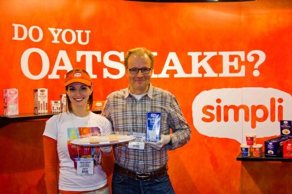 A woman in an orange visor holds a tray of small sample cups and a man in a plaid shirt holds a carton of oat drink, in front of an orange simpli backdrop reading "Do you oatshake?"