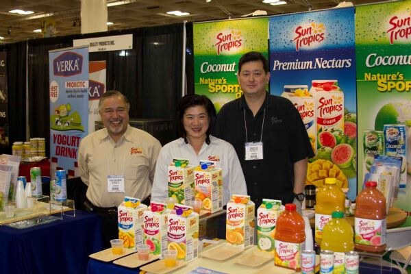 Two men and a woman stand behind a table of Sun Tropics juice cartons and bottles with sample cups. Sun Tropics banners and a Verka yogurt drink banner appear behind them.