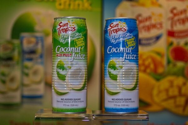 Two cans of Sun Tropics Hydration Coconut Juice, one with pulp in green and one in blue, stand on clear stands in front of blurred Sun Tropics packaging.