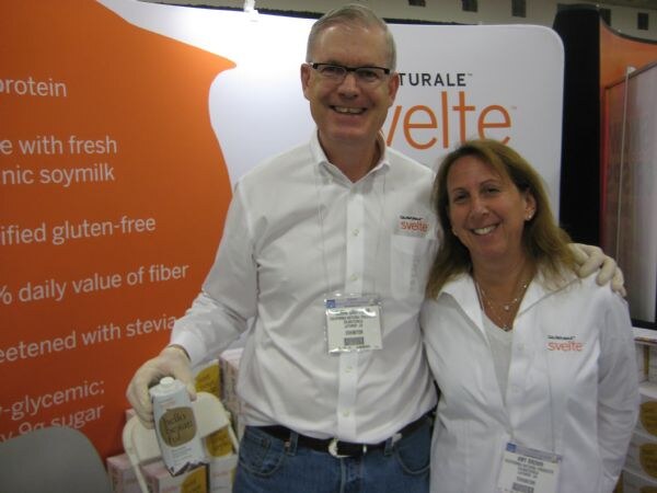 A smiling man with glasses holding a Svelte carton and a smiling woman, both in white shirts with Svelte logos and badges, stand in front of an orange Svelte display.