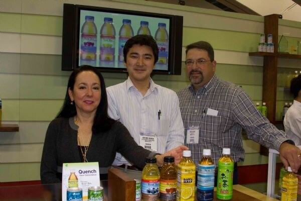 A woman and two men stand behind a counter lined with bottled Tea's Tea drinks and cans, below a screen showing bottles. The men wear badges.