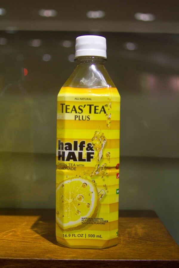 A yellow 16.9 oz bottle of Teas' Tea Plus half & half green tea with lemonade, with a lemon slice on the label, stands on a wooden surface against a dark background.