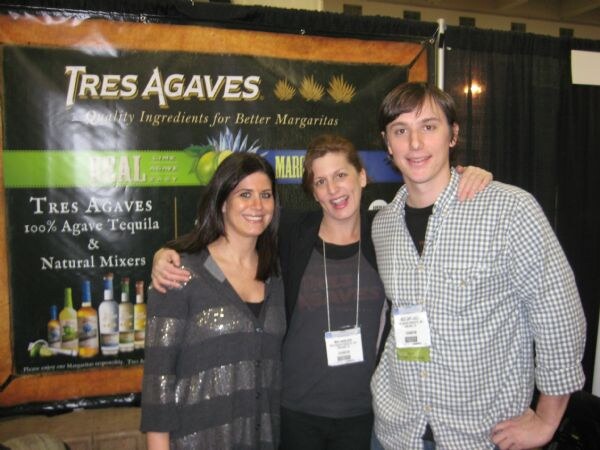 Three smiling people pose close together in front of a Tres Agaves banner advertising 100% agave tequila and natural mixers, with bottles shown on the banner.