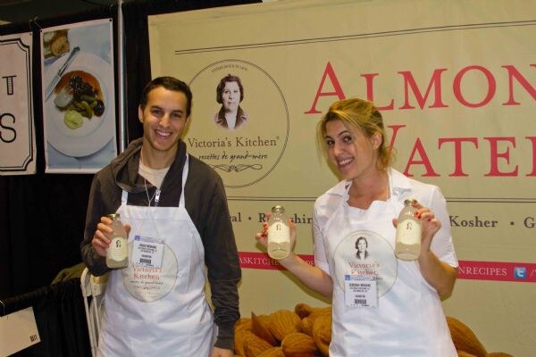 A man and a woman in white Victoria's Kitchen aprons smile and hold small bottles in front of an Almond Water banner, with a pile of almonds on the table.