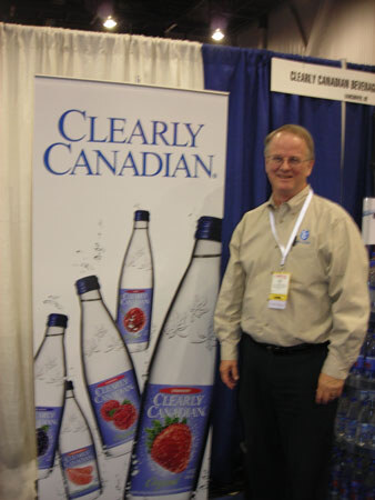 A smiling man in a tan shirt with a lanyard stands next to a large Clearly Canadian banner showing bottles of flavored sparkling water, including a strawberry flavor.