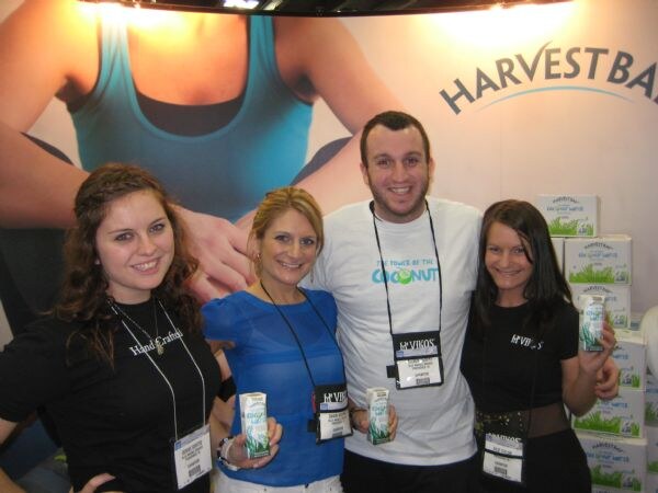 Four smiling people, three women and a man, wear badges and hold small cartons of Vikos coconut water. Cases of coconut water and a Harvest Bay sign are behind them.