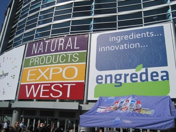 Large Natural Products Expo West and Engredea banners hang on a glass building above a blue Lifeway tent displaying bottles of kefir, with a crowd gathered below.