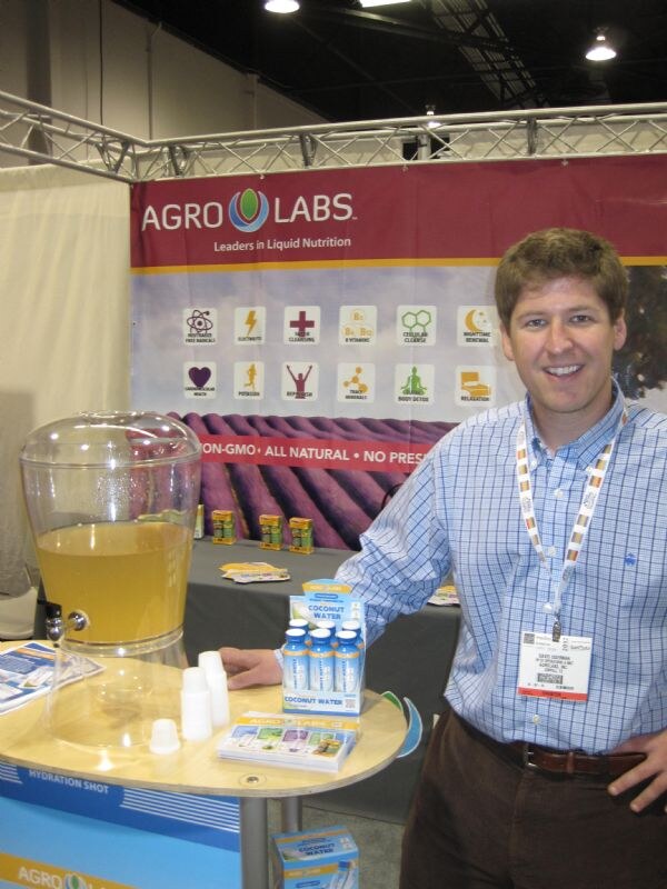 A smiling man in a blue checked shirt and lanyard stands at the Agro Labs booth beside a beverage dispenser and a display of coconut water hydration shots.