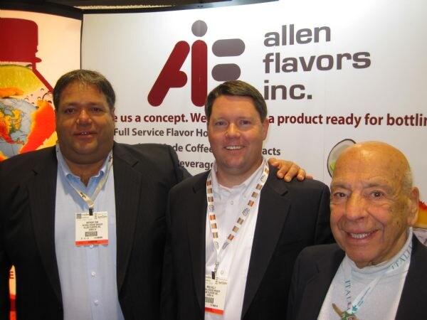 Three men in dark jackets pose closely together in front of an Allen Flavors Inc. banner; two wear show badges and lanyards.