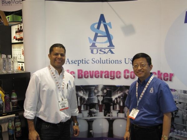 Two men in front of an Aseptic Solutions USA banner; one wears a white shirt and the other a blue shirt and glasses, both with badges and lanyards.