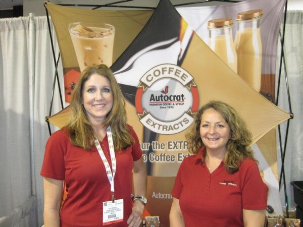 Two smiling women in red polo shirts stand in front of an Autocrat Coffee Extracts banner showing an iced coffee drink and bottles.