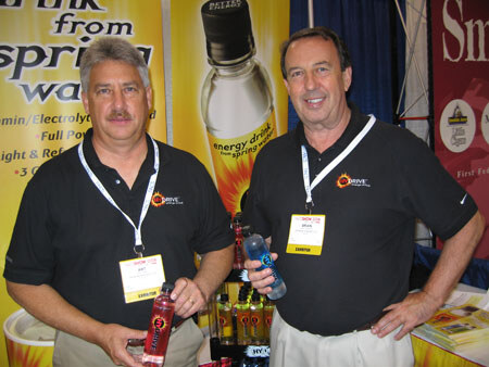 Two men in black Drive polo shirts with exhibitor badges each hold a bottle in front of Drive energy drink banners.