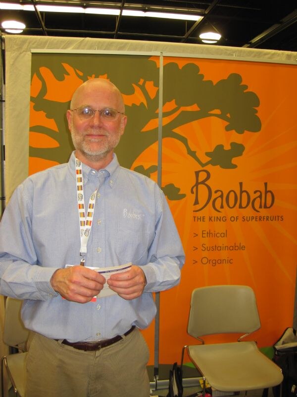 A bald, bearded man with glasses and a lanyard stands in front of an orange Baobab banner reading The King of Superfruits: Ethical, Sustainable, Organic.