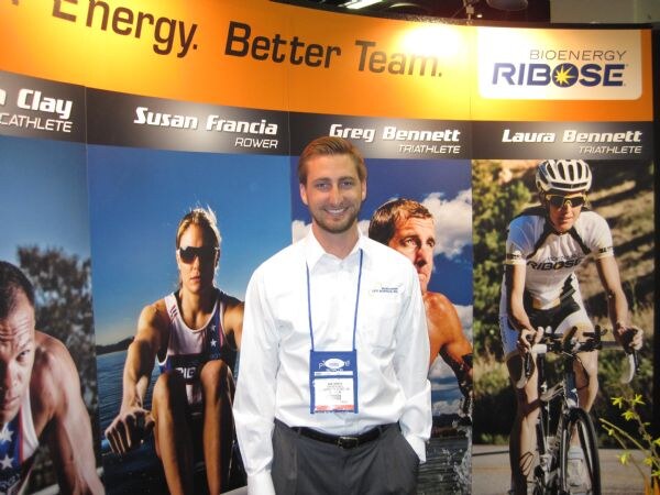 A smiling man in a white shirt and lanyard stands in front of Bioenergy Ribose banners showing athletes, including a rower and a cyclist.