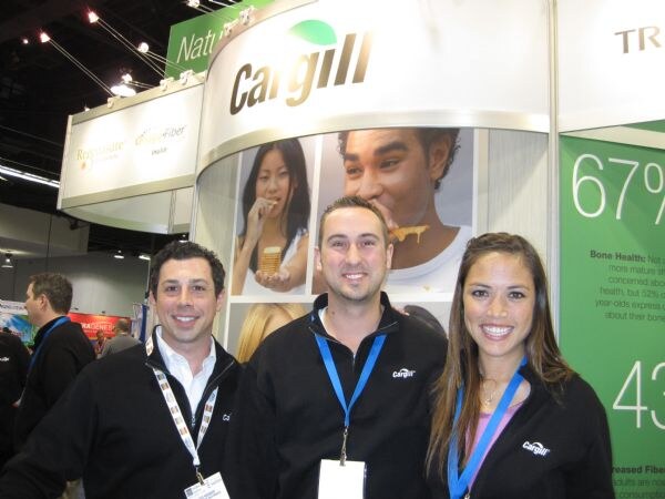 Two men and a woman in black Cargill jackets with lanyards smile in front of the curved Cargill booth sign and photos of people eating, beside a green panel showing 67%.