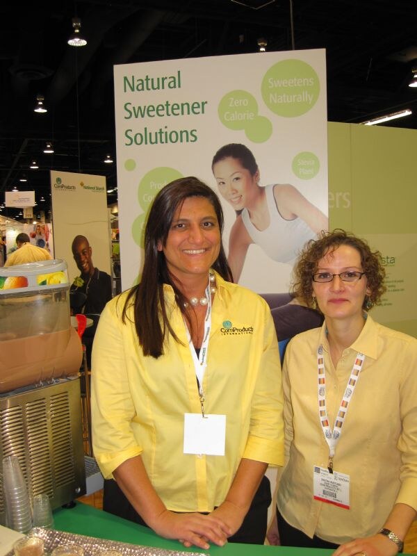 Two women in yellow shirts stand at a green counter in front of a Natural Sweetener Solutions banner; the woman on the left wears a Corn Products International logo shirt.