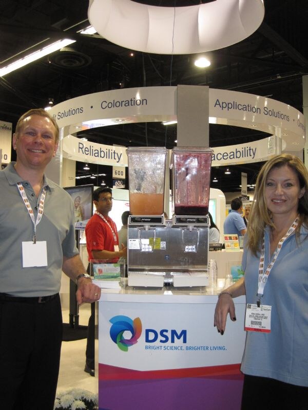 A man in a gray polo and a woman in a light blue polo stand beside a DSM counter holding two beverage dispensers, under a ring-shaped sign reading Reliability and Application Solutions.