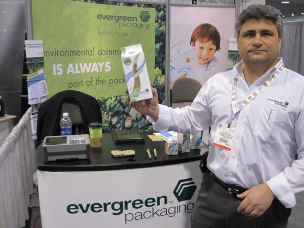 A man in a white Evergreen Packaging shirt holds up a carton beside a booth counter bearing the evergreen packaging logo, with a banner showing a smiling child behind him.