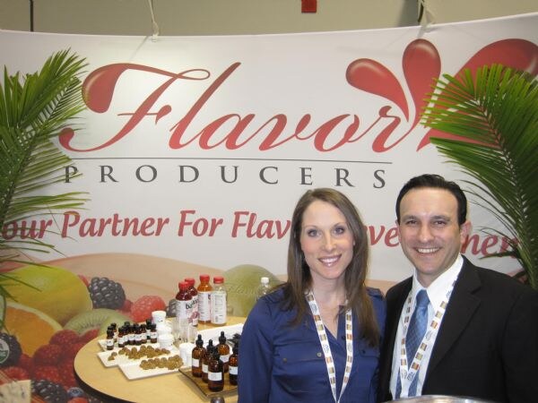 A woman in a blue top and a man in a dark suit smile in front of a Flavor Producers banner, with a table of small bottles and ingredient samples and palm fronds beside them.