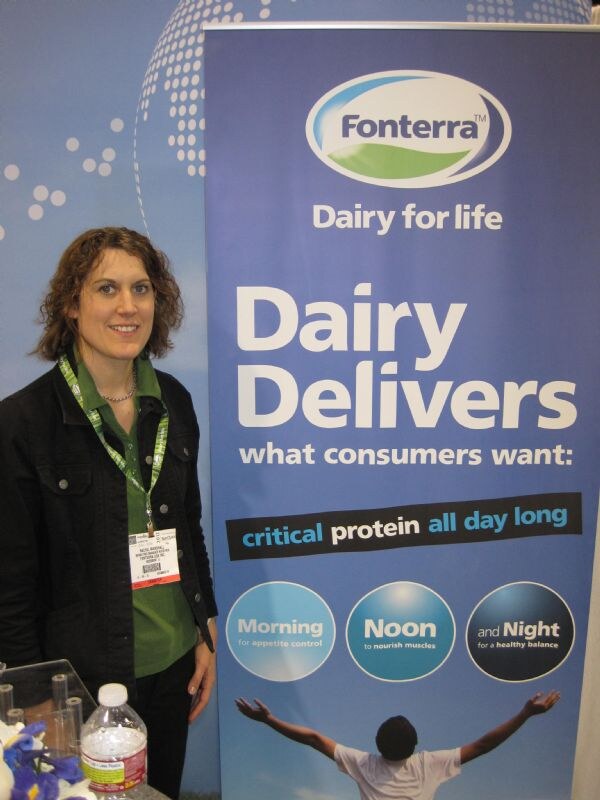 A woman with curly hair in a green shirt and black jacket stands beside a tall Fonterra banner reading Dairy Delivers what consumers want: critical protein all day long.
