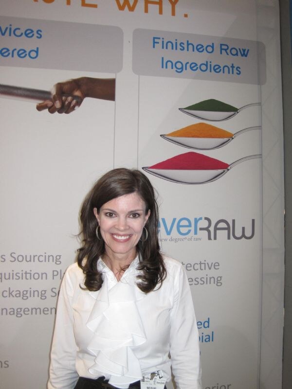 A smiling woman with long brown hair in a white ruffled blouse stands in front of an everRAW banner showing spoons of green, orange and red powders under Finished Raw Ingredients.