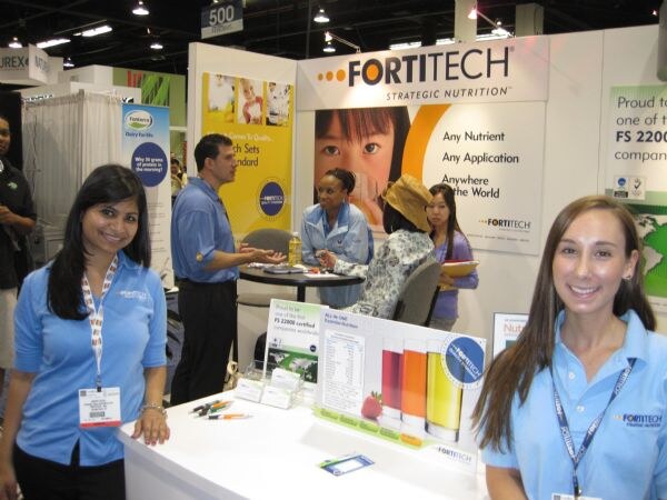 Two women in light blue Fortitech polos stand in the foreground of the Fortitech Strategic Nutrition booth, while other people talk at a table behind them.