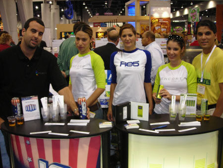 Five people stand behind two round display tables with product samples and cups. Several wear white shirts with green sleeves reading Encore or Re-5.