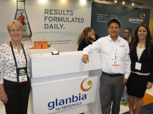 Two women and a man stand around a white Glanbia Nutritionals counter in front of signs reading Results Formulated Daily, with other people visible in the booth behind them.