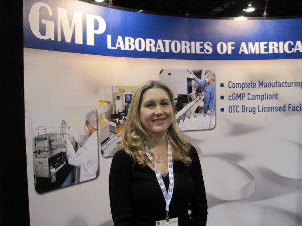 A smiling woman with long blonde hair and a lanyard stands in front of the GMP Laboratories of America booth, which has photos of lab workers and text on cGMP compliance.